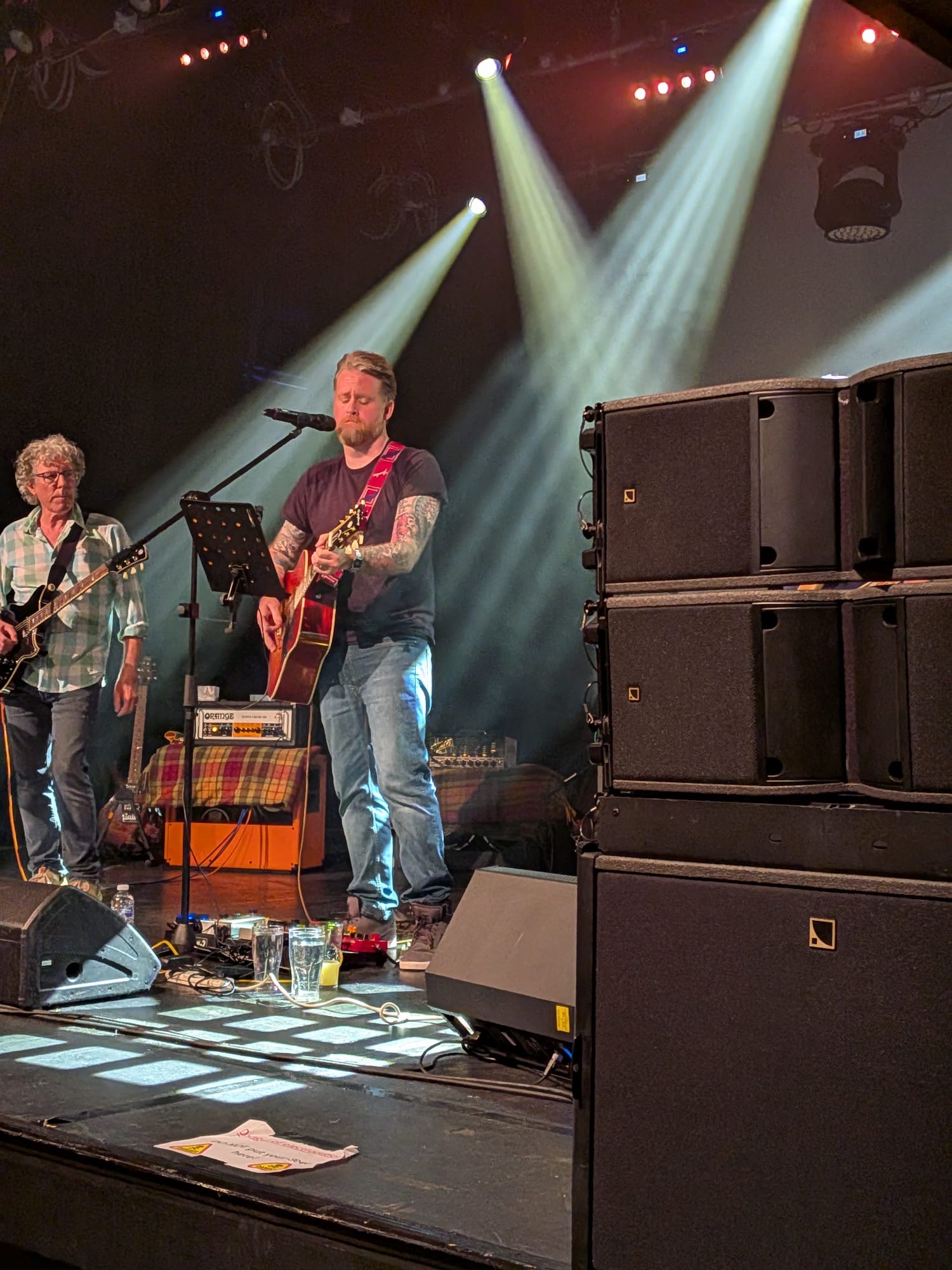 Two musicians performing on stage under white spotlights, one playing acoustic guitar at a mic, with a stacked line array PA to the side.
