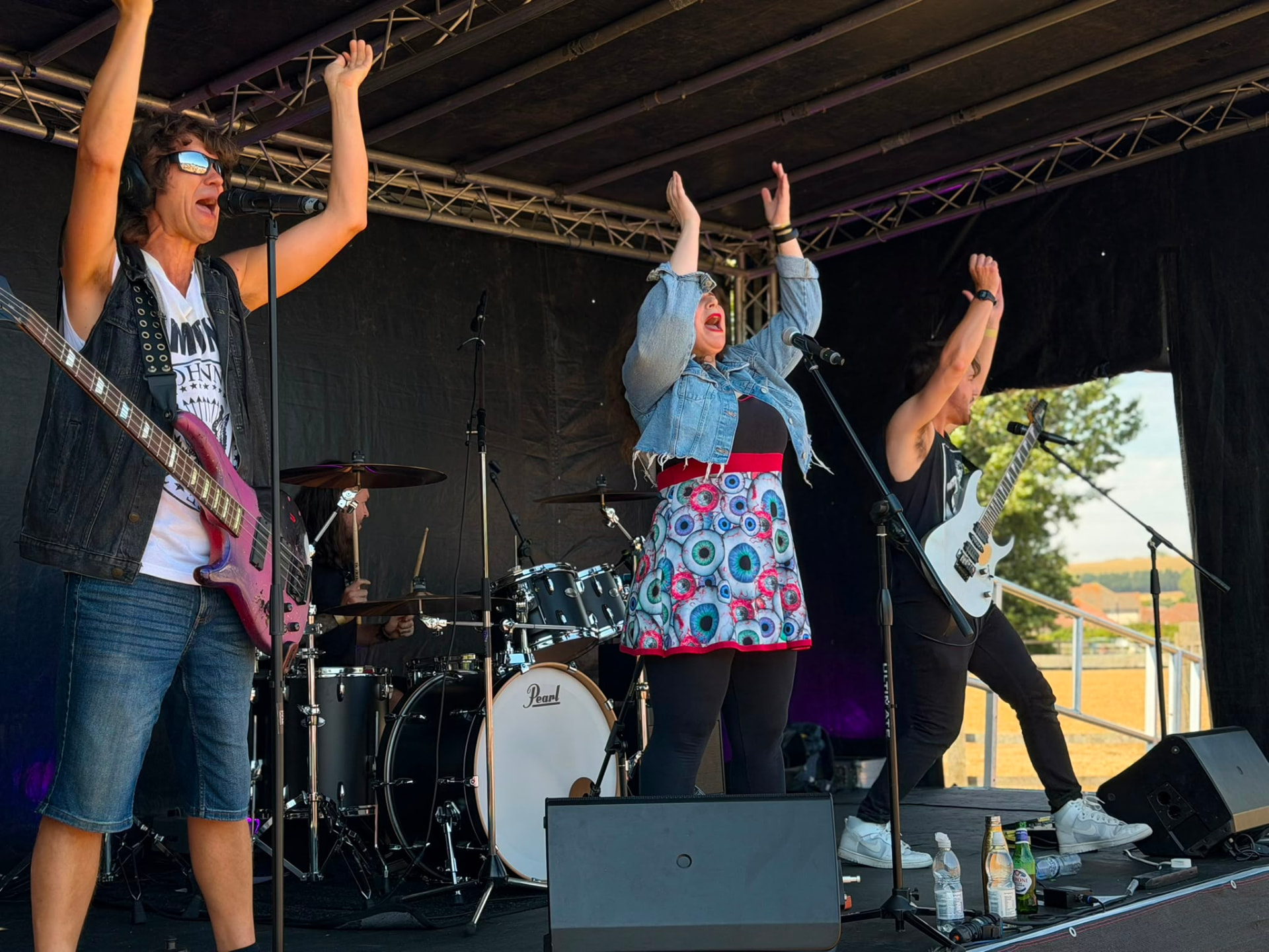A four-piece rock band performing on an outdoor stage with arms raised, bassist and guitarist either side of a singer and drummer.