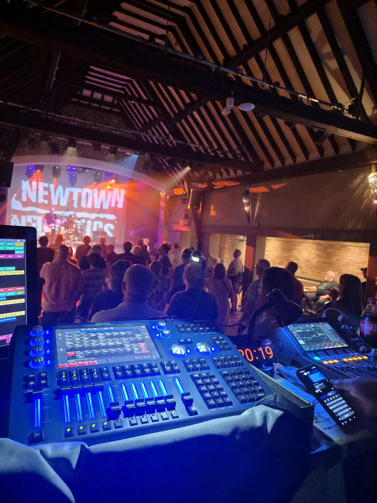 A lighting console and digital mixing desk at front of house, looking out over a busy crowd towards a band on a lit stage in a beamed barn venue.