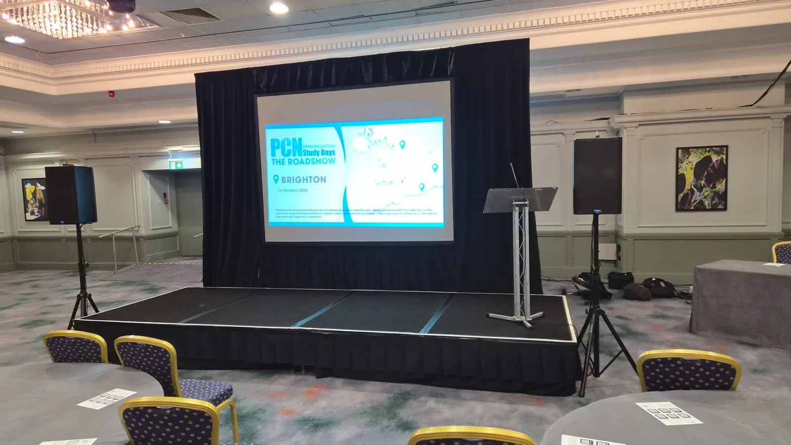 A conference room with a low stage, a projection screen framed by black drape, a lectern and speakers on stands either side.
