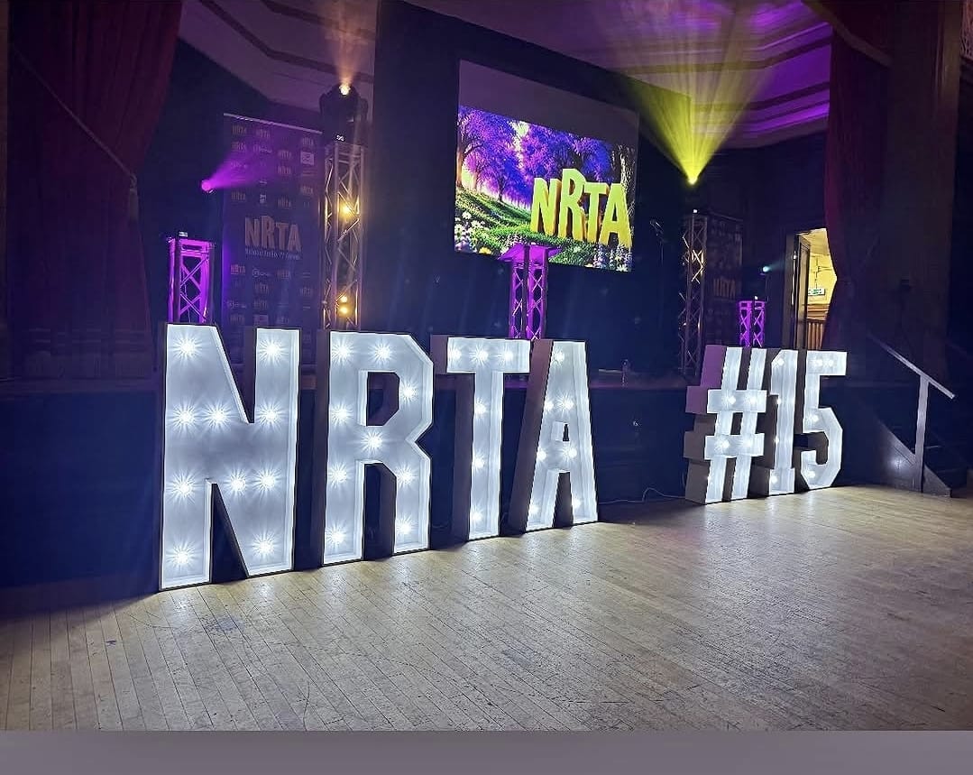Large illuminated NRTA #15 marquee letters in front of an awards stage with truss, projection screen and purple lighting.