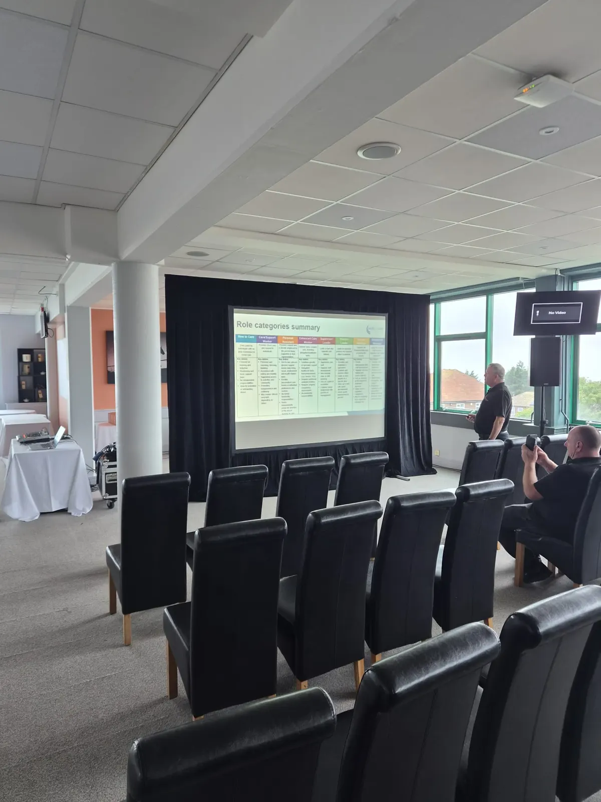 Rows of black chairs facing a projection screen framed in drape in a bright function room, with a speaker on a stand beside it.