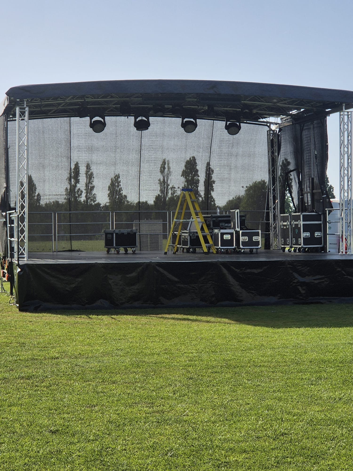 A covered outdoor stage on grass being set up, with overhead lighting, safety railing and black flight cases on the deck.