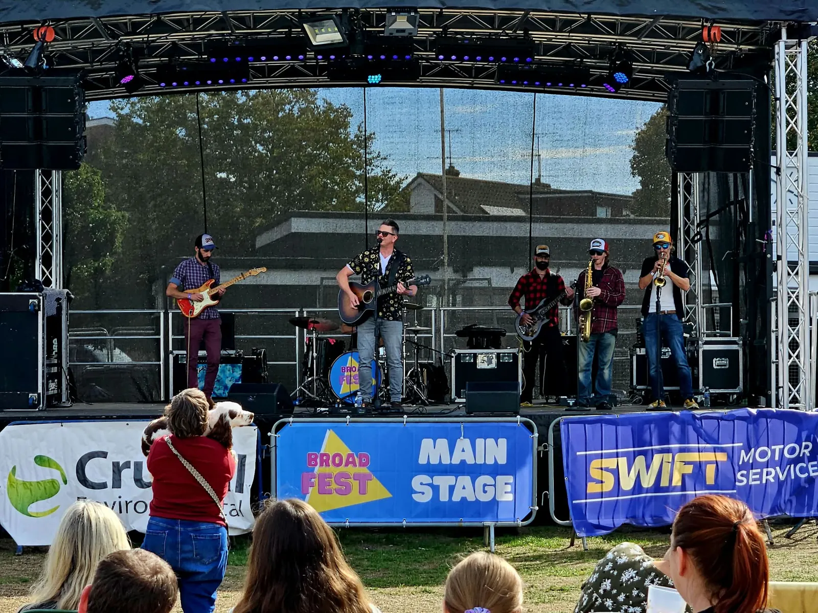 A band of guitarists performing on an outdoor main stage with Broadfest Main Stage banners along the barrier and an audience in front.