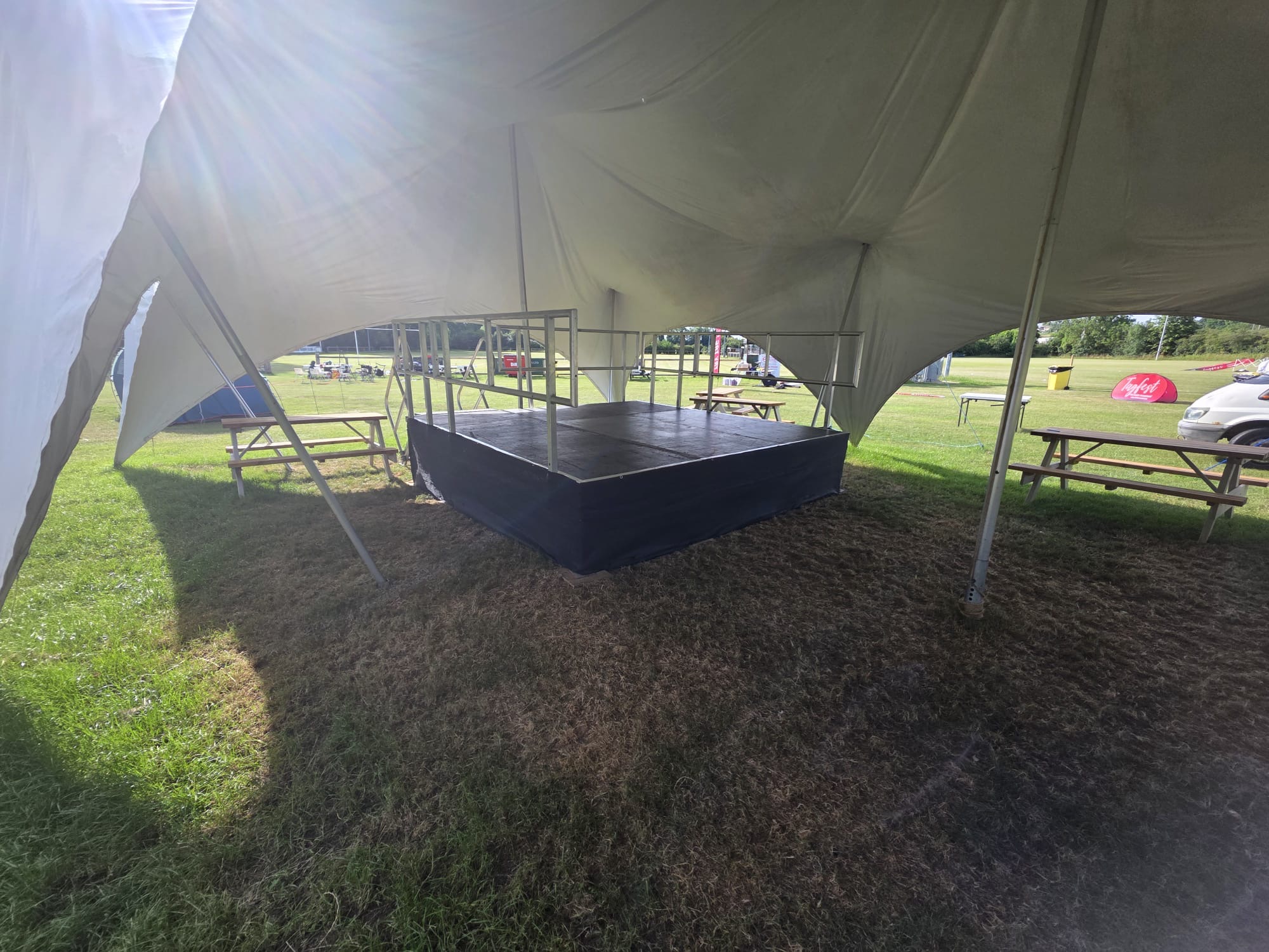 A low black stage with safety railing set up on grass under a marquee, with picnic benches either side and a sports field behind.