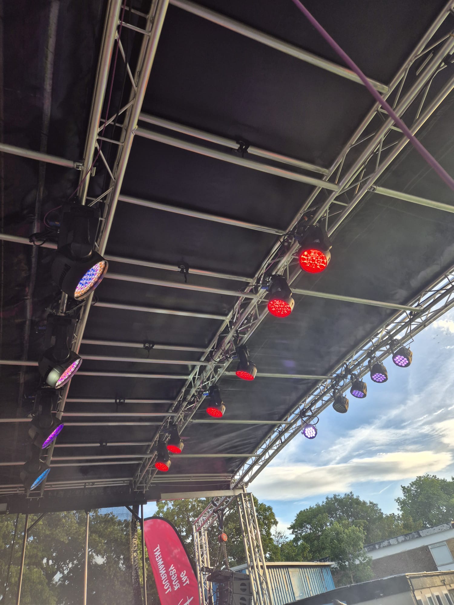 A close view up into a stage roof truss with rows of moving-head lights in red, purple and blue, with a 'Tag Rugby Tournament' banner below and blue sky beyond.