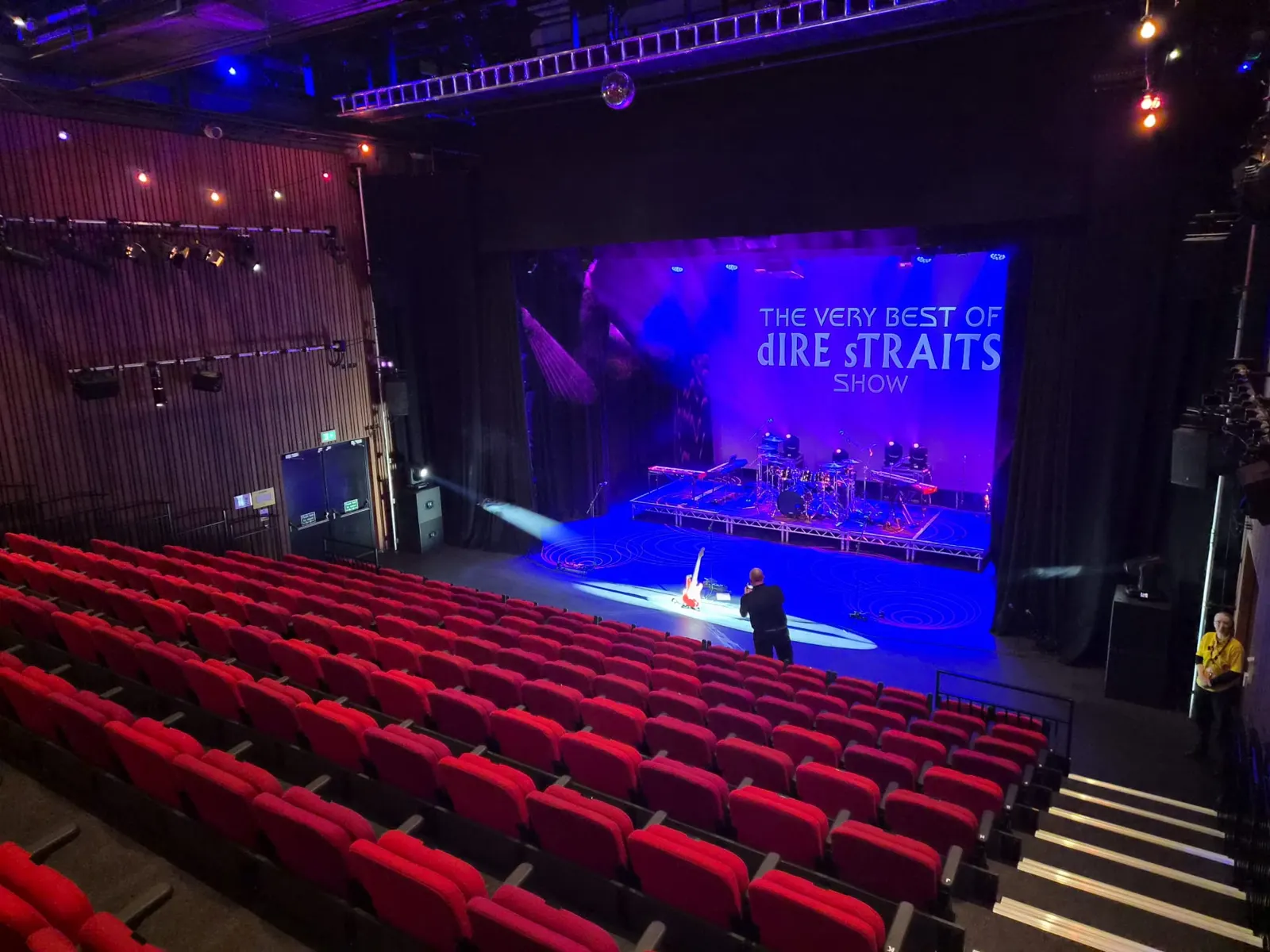 A theatre auditorium of red seats facing a stage lit in blue and purple, with a large screen reading The Very Best of Dire Straits Show.