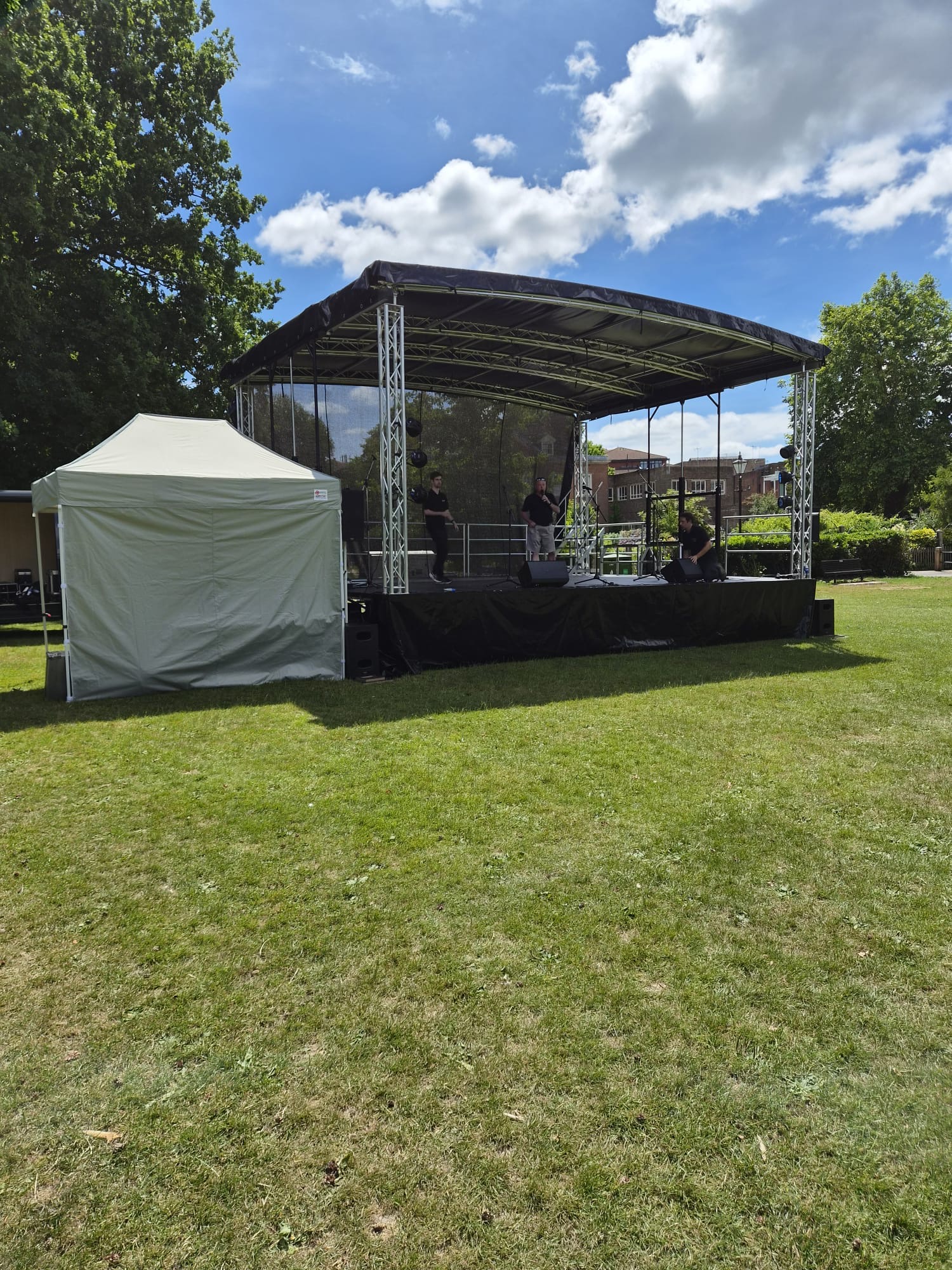A covered outdoor stage set up on grass in a park, with crew on stage and a small white marquee beside it under a blue sky.